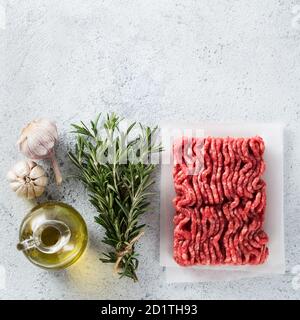 Bœuf haché cru frais, romarin frais, ail, huile d'olive sur fond de ciment gris clair avec espace de copie. Vue de dessus ou plan de travail. Concept d'ingrédients de cuisine. Forme carrée Banque D'Images