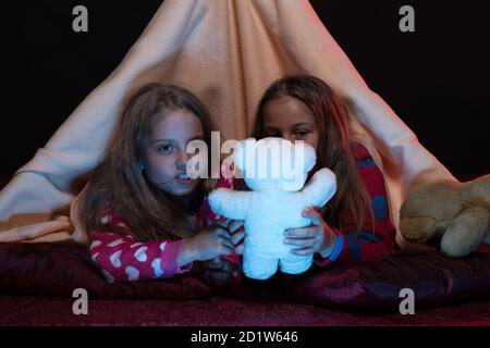 Pjs fête pour les enfants. Enfants et divertissement. Amis s'amusant avec des jouets dans une tente à couverture. Filles avec des visages heureux jouant avec des teddies. Les enfants portent des gambies rouges au lit sur fond noir. Banque D'Images