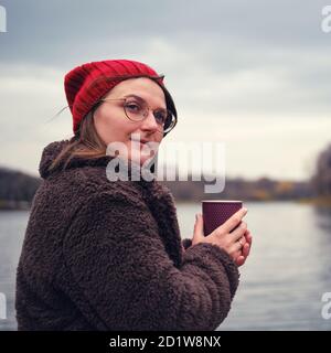 Une jeune femme se tient avec une tasse de café dessus la rive d'un étang d'automne Banque D'Images