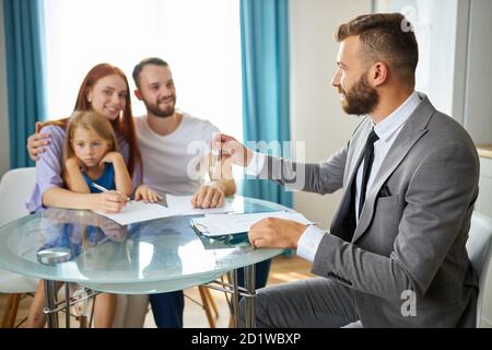 jeune famille caucasienne obtenant des clés de leur maison par agent immobilier, homme en costume leur donner des clés, famille heureuse avec enfant fille s'asseoir souriant Banque D'Images