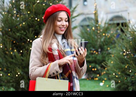 Portrait d'une jeune shopper souriante achetant en ligne sur un smartphone dans la rue de la ville avec des arbres de noël sur le fond Banque D'Images