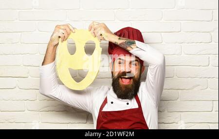 Faites cuire avec un visage excités dans des pièces d'uniforme bordeaux avec de la pâte à pizza. Concept du processus de cuisson. Le chef prépare de la pâte. Homme avec barbe tient la pâte roulée avec smiley coupé sur fond de mur de brique blanche. Banque D'Images