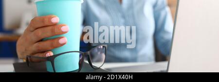 La main féminine tient le verre bleu avec le café sur le lieu de travail. Banque D'Images