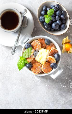 Bol avec de minuscules céréales pancakes sur un fond en béton léger. Cuisine tendance. Mini crêpes aux céréales avec beurre, miel et bleuets. Banque D'Images