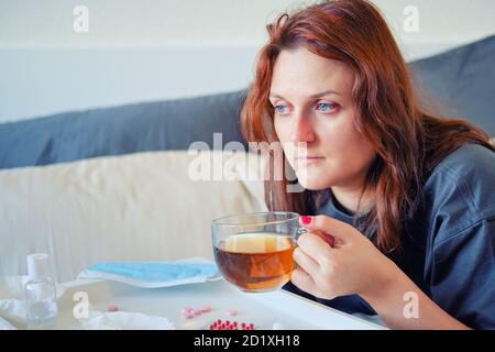 Une femme aux cheveux rouges avec le virus de la grippe boit du thé. Fille avec un coronavirus assis sur le lit Banque D'Images