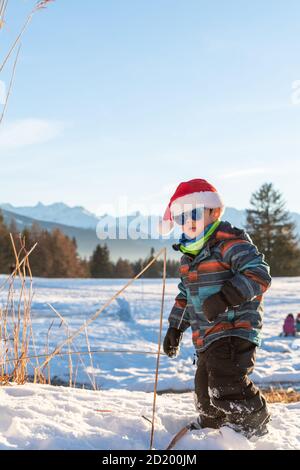 Garçon avec chapeau rouge de Santa sur fond de paysage d'hiver. Joyeux sourire enfant dans la neige à Noël debout à côté de grandes plantes à l'extérieur. Crans Banque D'Images