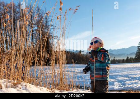 Garçon avec chapeau rouge de Santa sur fond de paysage d'hiver. Joyeux sourire enfant dans la neige à Noël debout à côté de grandes plantes à l'extérieur. Crans Banque D'Images