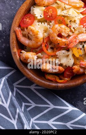 Pâtes farfalle aux crevettes, épinards et tomates cerises dans une assiette en bois, parsemées de poivre fraîchement moulu parfumé sur la table Banque D'Images