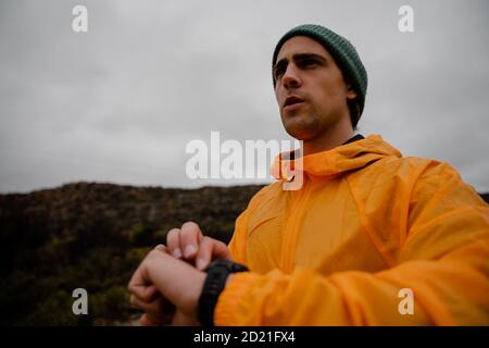 Belle tenue jeune homme randonneur contrôle de la fréquence cardiaque sur la montre intelligente en randonnée sur le sentier de montagne par temps nuageux Banque D'Images