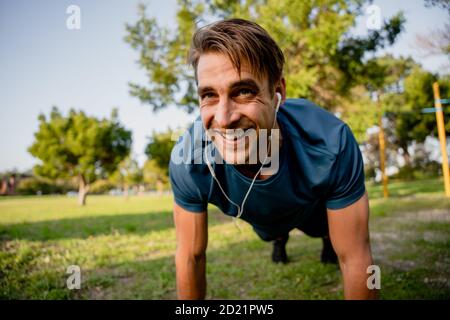 Souriant beau jeune athlète masculin planking exercice en plein air ensoleillé ecouteurs de stationnement pour écouter de la musique Banque D'Images