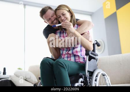 Le mari encadre sa femme en fauteuil roulant avec des épaules positives test de grossesse Banque D'Images