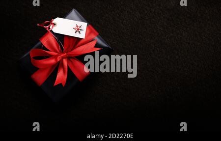 Un cadeau de Noël enveloppé de papier noir et de ruban rouge avec une étiquette vierge, carte sur un fond sombre par le dessus. Banque D'Images