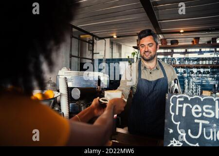 Une femme afro-américaine recueille du café auprès d'un serveur masculin souriant, debout derrière le comptoir dans un café élégant. Banque D'Images