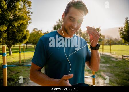 Jeune athlète masculin avec montre intelligente portant des écouteurs à l'écoute musique sur smartphone pendant l'entraînement dans le parc extérieur Banque D'Images