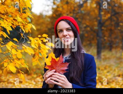 Souriante femme brune dans un chapeau rouge tenant des feuilles d'érable dans les deux mains. Concept d'automne. Banque D'Images