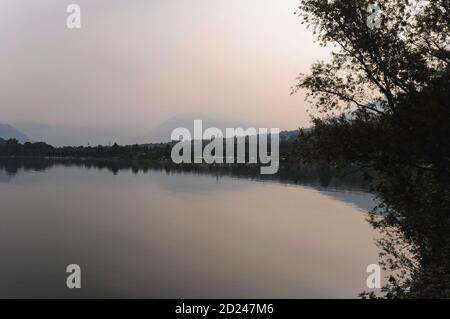 La brume matinale obscurcit les sommets alpins lointains du canton suisse du Tessin, de langue italienne, bien au-delà de la petite ville de Laghetto et des eaux calmes du Laghetto di Piona, une baie tranquille, populaire auprès des baigneurs, sur la rive nord-est du Lago di Como en Lombardie, en Italie. Les points d'observation en bord de route sur la baie offrent également une vue superbe sur le bras nord du lac de Côme vers les colonies de ses rives nord-ouest, y compris la ville historique de Gravedona. Banque D'Images