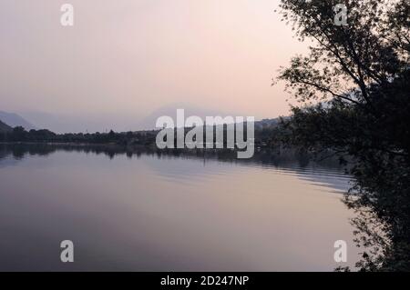 La brume matinale obscurcit les sommets alpins lointains du canton suisse du Tessin, de langue italienne, bien au-delà de la petite ville de Laghetto et des eaux calmes du Laghetto di Piona, une baie tranquille, populaire auprès des baigneurs, sur la rive nord-est du Lago di Como en Lombardie, en Italie. Les points d'observation en bord de route sur la baie offrent également une vue superbe sur le bras nord du lac de Côme vers les colonies de ses rives nord-ouest, y compris la ville historique de Gravedona. Banque D'Images