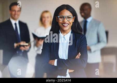 Une jeune femme d'affaires africaine debout avec ses bras a traversé une bureau et sourire avec des collègues debout en arrière-plan Banque D'Images