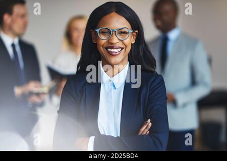 Jeune femme d'affaires africaine souriante debout dans un bureau avec un divers groupes de collègues se tenant en arrière-plan Banque D'Images
