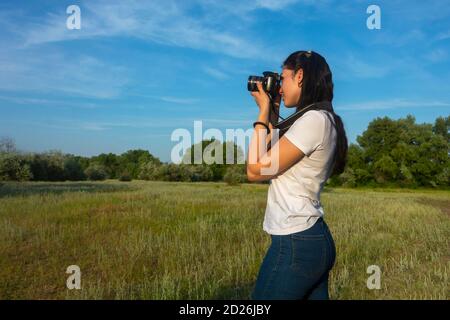 La jeune femme photographie le paysage en été. Vue latérale d'un magnifique brunette tenant un appareil photo numérique. Magnifique ciel bleu et forêt autour. Banque D'Images