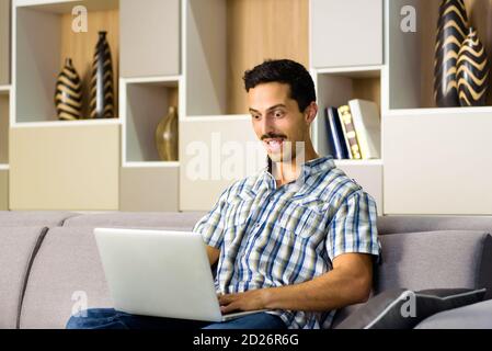 Un jeune homme réagit avec étonnement à un ordinateur portable qui est sous la féérie large-oculaire avec sa bouche agape comme il se détend à la maison sur un canapé confortable Banque D'Images