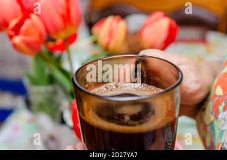 Femme âgée tenant une tasse de verre noir de café chaud buvez sur un fond de tulipes rouges sur la table Banque D'Images