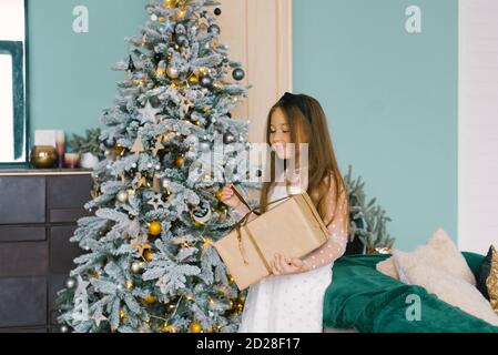 Bonne fille belle ouvre un cadeau de Noël tout en se tenant près Un sapin de Noël festif Banque D'Images