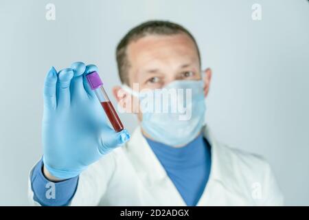 Un médecin masculin dans une robe médicale, un masque et des gants tient un tube à essai avec du sang. Concept de tests médicaux Banque D'Images