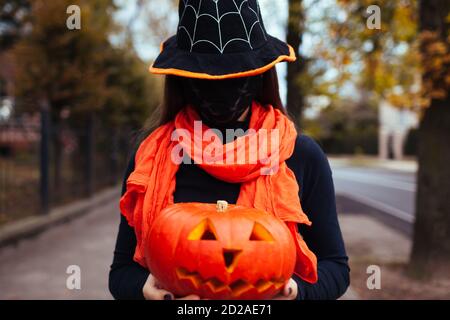 Citrouille en forme de citrouille d'Halloween. Effrayante fille tient la citrouille sculptée dehors portant le masque contre le coronavirus cachant visage. Banque D'Images