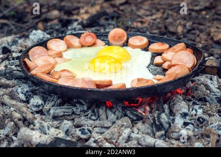 Deux œufs et saucisses sont frits dans une poêle sur des charbons dans la forêt. Petit déjeuner dans la nature, pique-nique. Banque D'Images