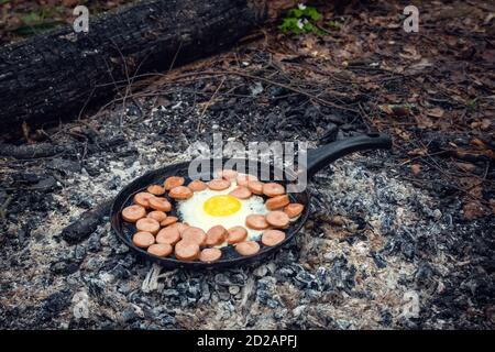 les œufs et les saucisses sont frits dans une poêle sur des charbons dans la forêt. Petit déjeuner dans la nature, pique-nique. Banque D'Images