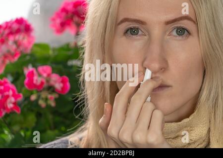 Jeune femme blonde utilise un spray nasal pour un écoulement nasal ou allergie sur un fond de fleurs intérieures, espace de copie. Banque D'Images