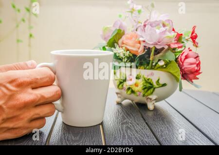 La main d'un homme tient une tasse de café blanc sur une table en bois près, à côté d'un bouquet de fleurs Banque D'Images