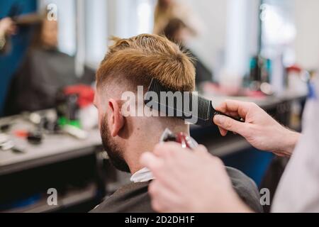 Gros plan sur un homme qui obtient une coupe de cheveux tendance au salon de coiffure. Coiffeur masculin servant le client, faisant la coupe de cheveux à l'aide de la machine et du peigne Banque D'Images