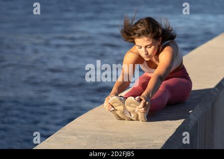 Jeune coureur de yoga femme en legging rose assis sur un remblai, étirant les muscles en position assise courbe vers l'avant. Exercices d'échauffement en stretch avec havotre Banque D'Images