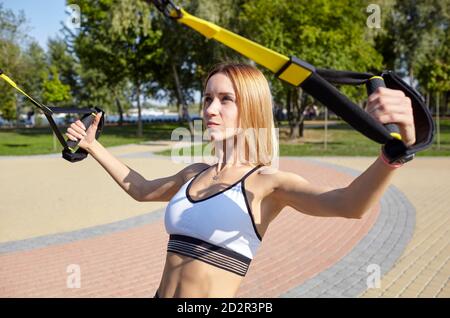 Effectuer des exercices TRX. Jeune femme athlétique avec corps parfait dans les vêtements de sport d'entraînement ses bras avec trx de forme dans le parc de la ville Banque D'Images