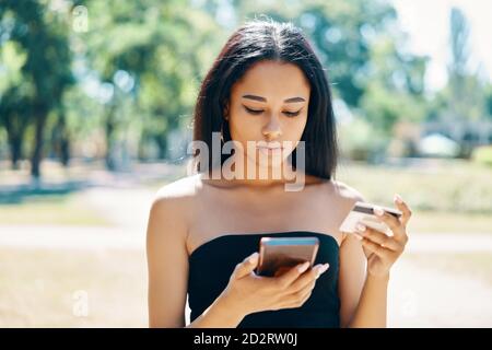 Femme noire payant avec une carte de crédit sur un smartphone à l'extérieur marche dans le parc Banque D'Images