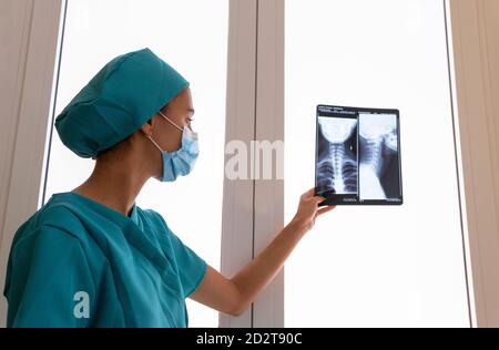 Vue latérale d'une jeune femme médecin spécialiste en blouse chirurgicale Et masque de protection examinant l'image radiographique du cou cervical colonne vertébrale du patient à l'hôpital Banque D'Images