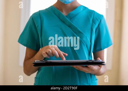 Jeune infirmière en gommages bleus et informations de lecture de masque médical sur une tablette dans un couloir de l'hôpital moderne Banque D'Images