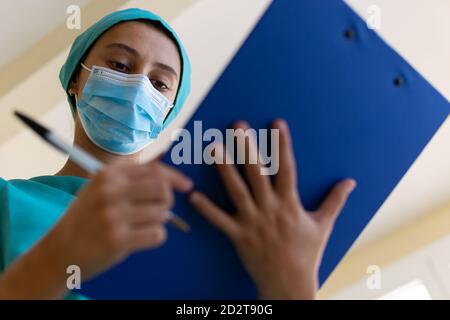 Jeune infirmière concentrée en uniforme bleu et masque faisant médical notes sur le presse-papiers pendant le travail à l'hôpital Banque D'Images