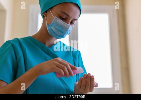 Faible angle de jeune infirmière dans les exfoliations bleues et médicales masquer l'application de l'assainisseur sur les mains lors de la préparation de la procédure dans clinique Banque D'Images
