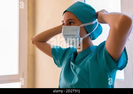 Vue latérale d'une infirmière féminine sérieuse en uniforme masque médical avant le travail en se tenant en clinique pendant le coronavirus épidémie et regarder loin Banque D'Images