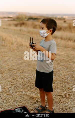 Petit garçon asiatique dans un masque de protection utilisant la télécommande pendant jouer avec un drone volant sur le terrain Banque D'Images