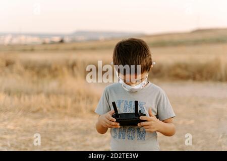 Petit garçon asiatique concentré dans un masque de protection à l'aide de la télécommande en jouant avec un drone volant sur le terrain Banque D'Images