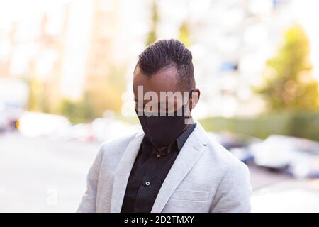 Un homme d'affaires afro-américain sérieux en costume formel et masque de protection debout dans la rue pendant la pandémie du coronavirus et en regardant vers le bas Banque D'Images