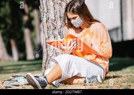 Jeune femme dans le masque de visage assis près du tronc d'arbre et de lire des notes dans le planificateur tout en faisant des devoirs le jour ensoleillé à Madrid, Espagne Banque D'Images