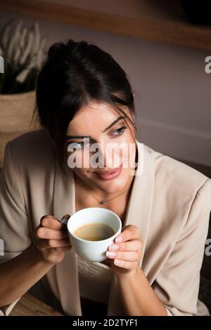 De dessus de positive jeune femme rêveuse avec une tasse de café aromatique assis à la table et donnant sur l'extérieur tout en se relaxant à la maison Banque D'Images