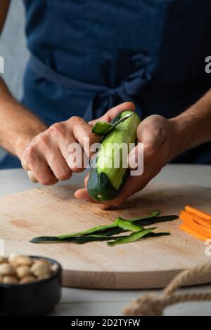 Croiser anonyme mâle cuire enlever peeling de la courgette verte fraîche tout en préparant un déjeuner sain dans la cuisine Banque D'Images