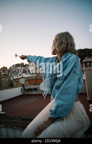 Vue latérale d'une femme rêveuse assise sur le toit du bâtiment et le sort de casting avec baguette magique dans la main tendue pendant vue sur l'extérieur Banque D'Images