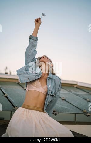 Vue latérale d'une femme rêveuse assise sur le toit du bâtiment et le sort de casting avec baguette magique dans la main tendue pendant recherche Banque D'Images